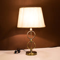 Table lamp with circular gold metal frame and beige fabric shade on white surface against brown wall.